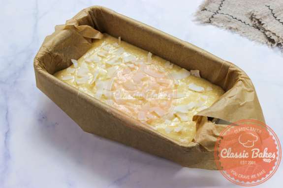 A loaf pan lined with parchment paper filled with batter topped with shredded coconut 