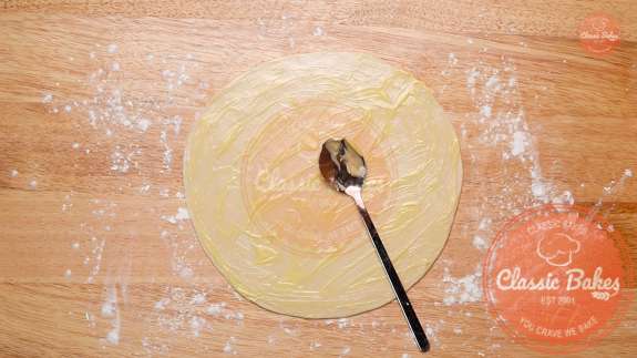 Circle of dough being rubbed with ghee using the back of a spoon 