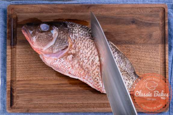 Overview of a whole snapper on a cutting board with a knife slicing the skin 