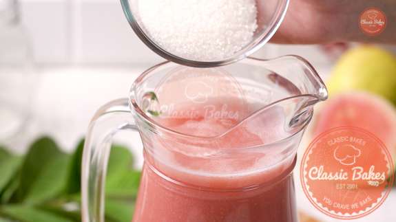 A small bowl of sugar being added to a pitcher of guava juice 