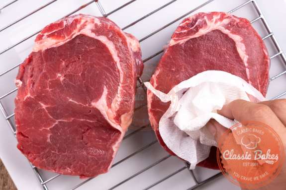Two ribeye steaks on a metal rack being patted dry with paper towel 