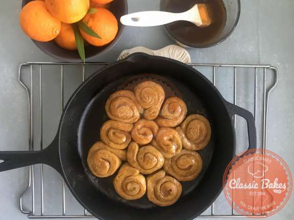 Overview of baked orange rolls in a skillet cooling on a rack next to a bowl of the butter and sugar mixture