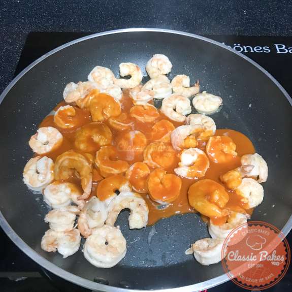Overview of buffalo sauce being added to a pan of cooked shrimp 
