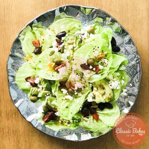 Overview of a platter of dressed butter lettuce garnished with tomatoes, olives, avocado, feta cheese and punpkin seeds  