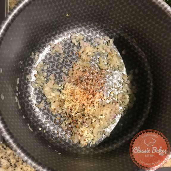Red pepper flakes being stirred into a pot of shallots and garlic cooking inside of it. 