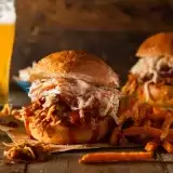 Sideview of two BBQ chicken sandwiches on a wooden board with fries and a glass of beer in the background 